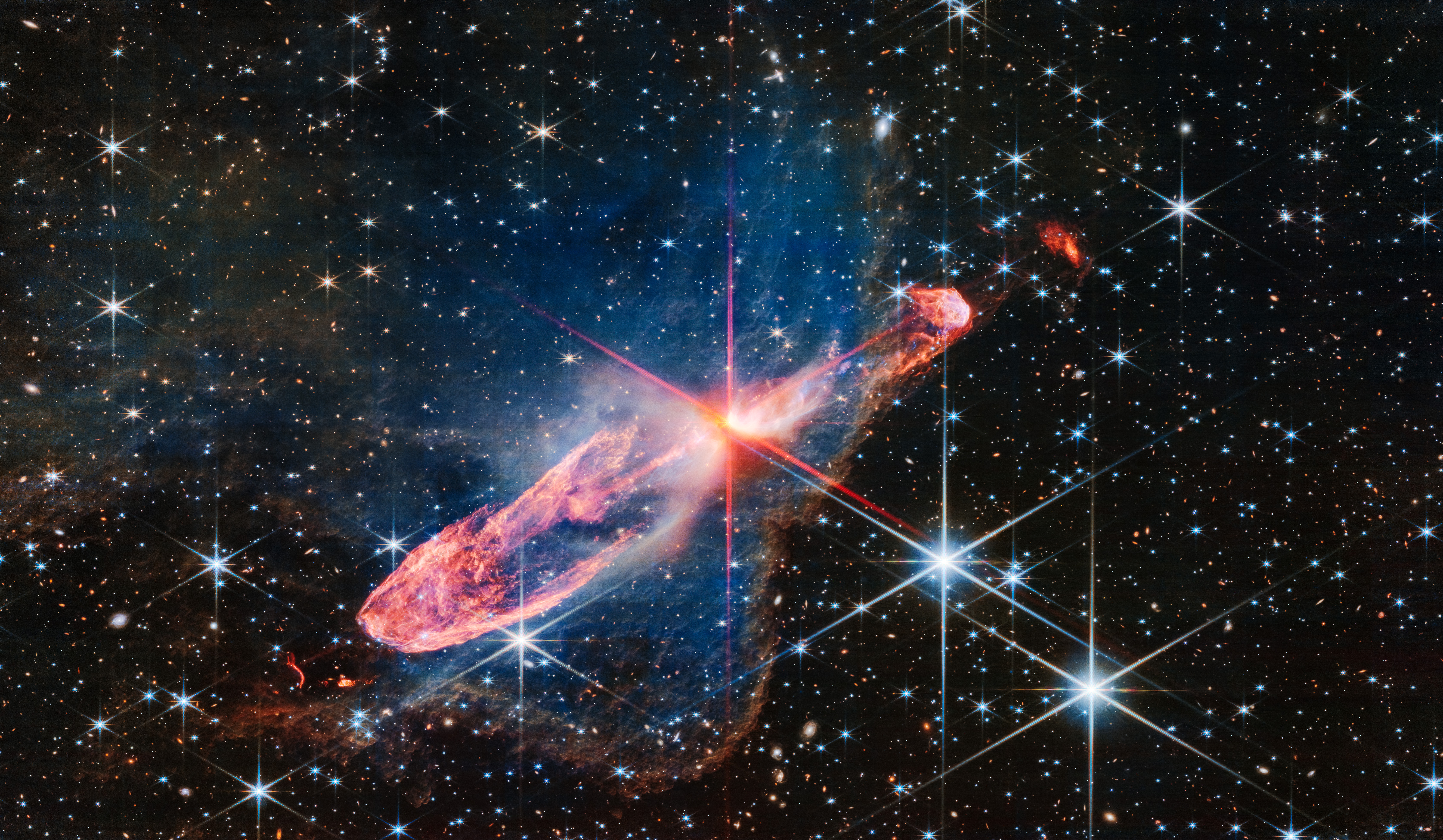 Horizontal orange cloud known as Herbig-Haro 46/47, with a central yellow-white blob pierced by red diffraction spikes. A surrounding nebula is seen as a delicate, semi-transparent blue haze. The background is filled with stars and galaxies. 